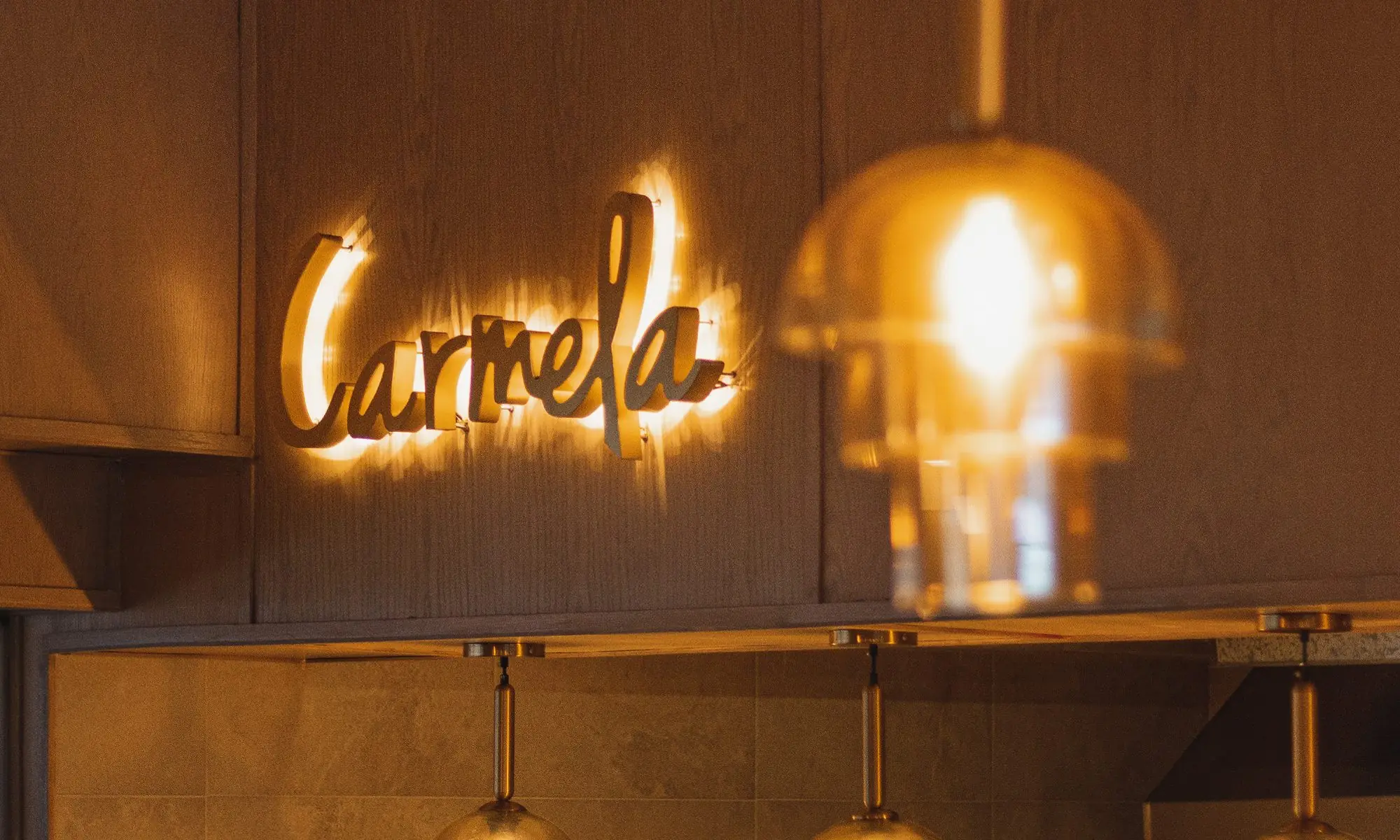 El letrero de Carmela iluminado sobre el muro de madera de Plaza Ceiba, con lámparas de cristal